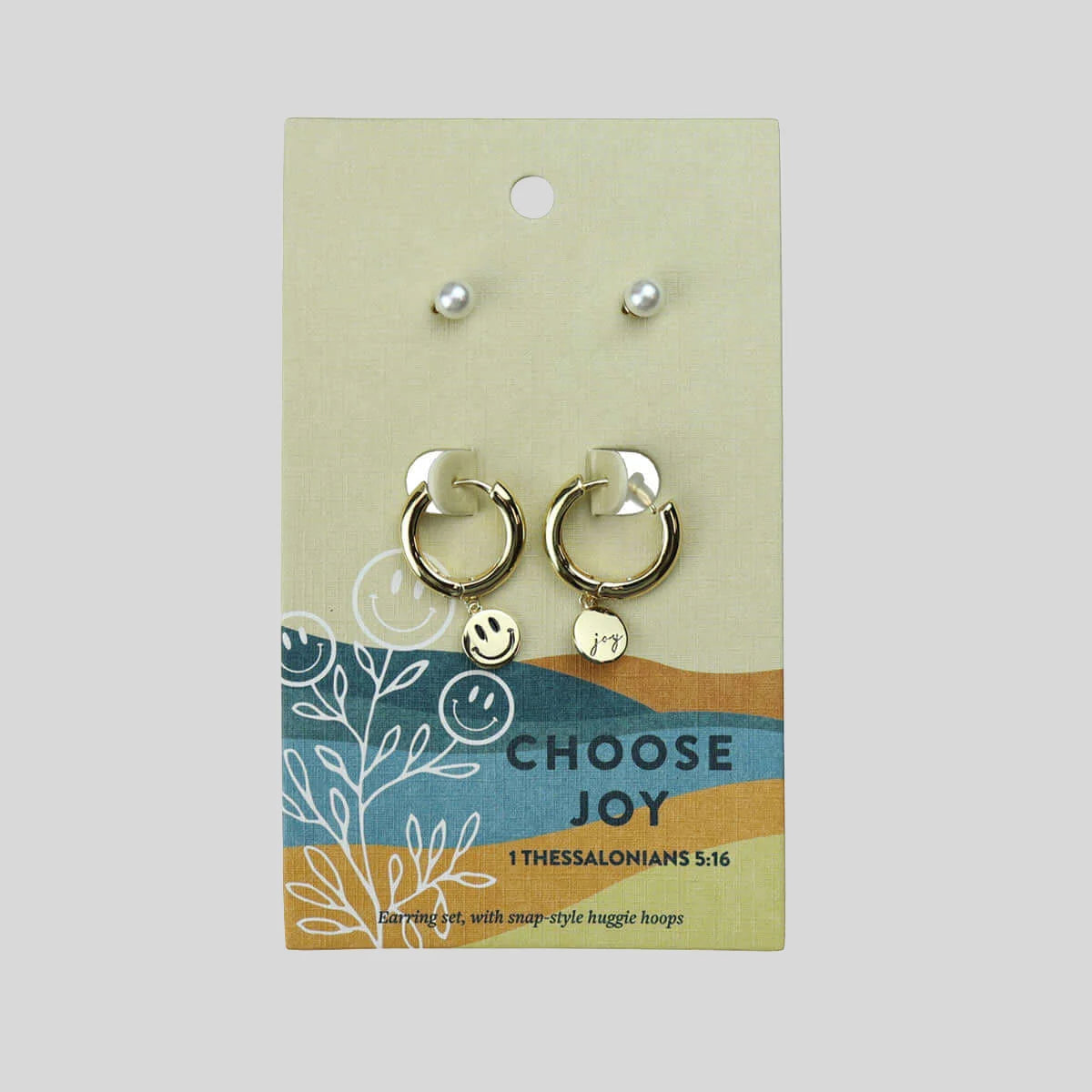 A set of earrings featuring a pair of gold hoop earrings with small smiley face charms and two pearl stud earrings above them, displayed on a beige card labeled 'CHOOSE JOY', against a light gray background.