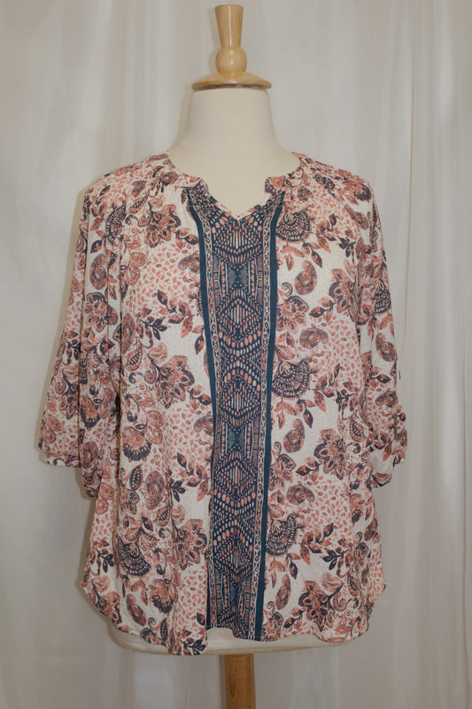 A women's bohemian-style blouse with a floral and paisley print in rose pink, white, and navy blue, featuring a central geometric patterned panel, displayed on a dress form.